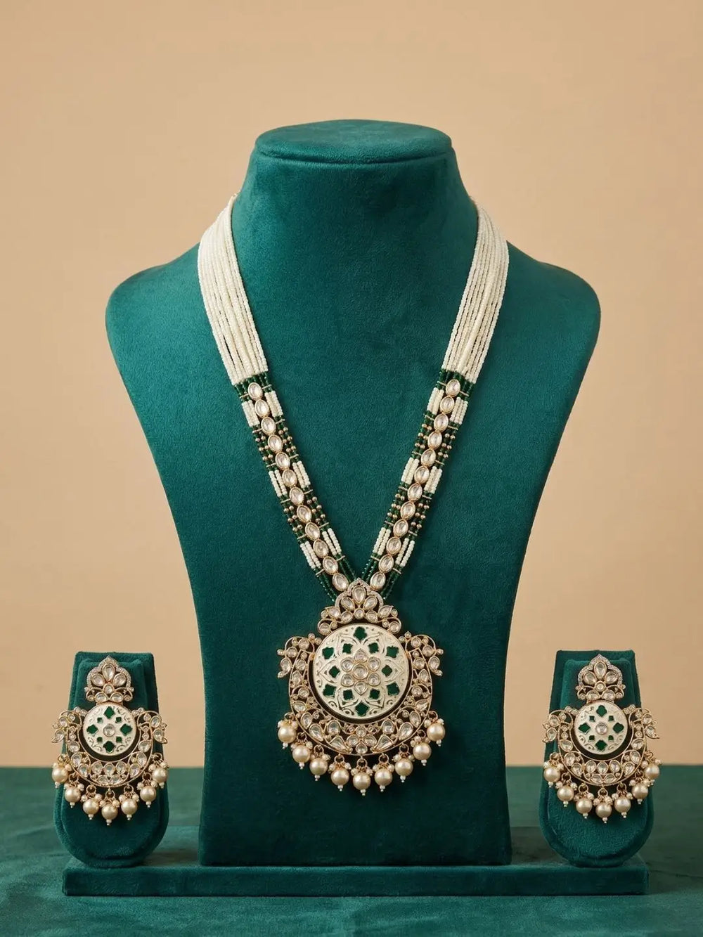 A traditional Indian jewelry set displayed on a dark teal velvet mannequin bust. The set includes a long necklace with a large ornate pendant, matching earrings, and a pair of bangles, all featuring white stones, green stones, and gold-toned metal.