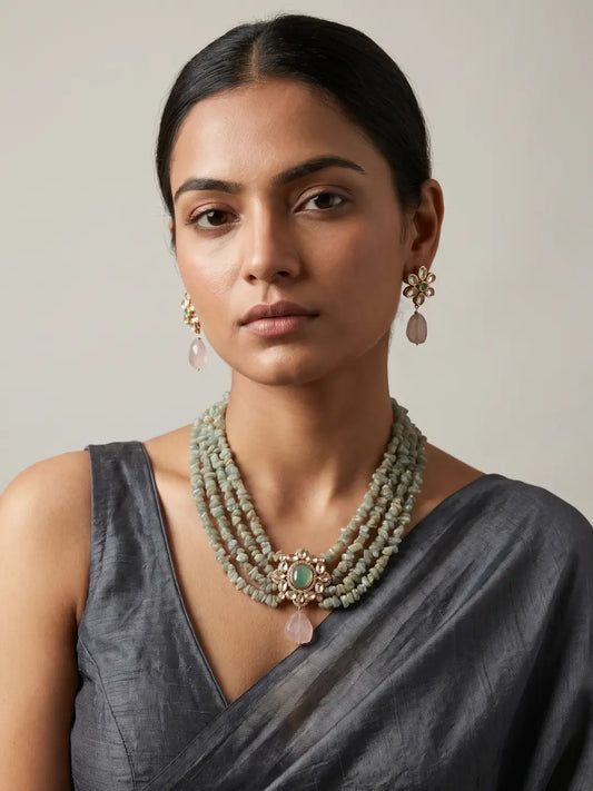A woman wearing a multi-strand beaded necklace with a central green stone and pink teardrop kundan pendant, matching drop earrings, and a dark gray sari against a neutral beige background.