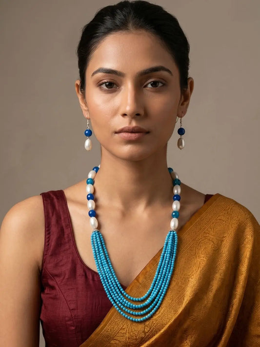 A woman wearing a multi-strand turquoise and white beaded necklace with matching drop earrings, styled with a maroon blouse and a gold saree against a neutral beige background.