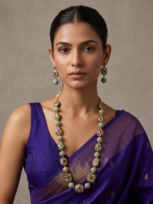 Woman wearing a long designer turquoise Jadau beads mala with multicolour antique metallic beads, green and ruby-red stone accents, gold-tone detailing and matching earrings.