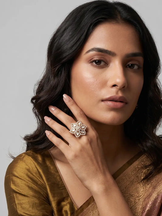 A close-up portrait of a woman wearing a large, ornate gold-toned polki ring with a floral or starburst design on one finger, set against a neutral gray background.