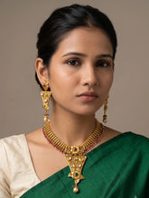 A woman wearing a traditional Indian gold-plated antique gold necklace and matching drop earrings, styled with a green saree and cream blouse against a neutral beige background.