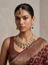 A woman wearing a traditional Indian outfit with a maroon and gold saree, styled with a gold-plated polki kundan necklace set with green stones and a matching earrings, and a maang tikka.