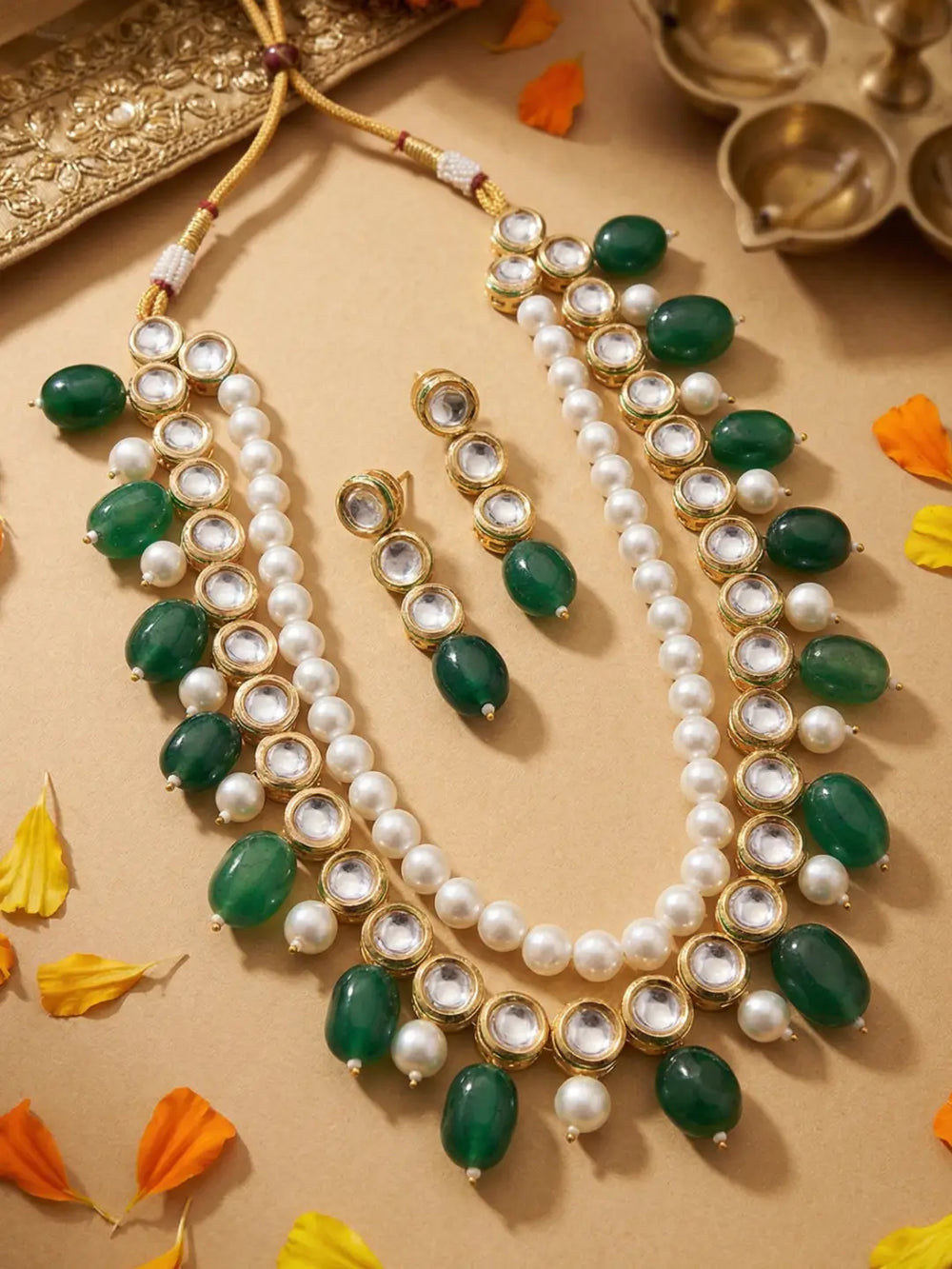 A gold-plated Indian kundan jewelry set featuring a multi-strand necklace with kundan string, green stone beads and white faux pearls, accompanied by matching drop earrings, arranged on a beige surface with decorative flower petals and traditional brass oil lamps.