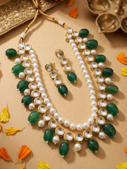 A gold-plated Indian kundan jewelry set featuring a multi-strand necklace with kundan string, green stone beads and white faux pearls, accompanied by matching drop earrings, arranged on a beige surface with decorative flower petals and traditional brass oil lamps.