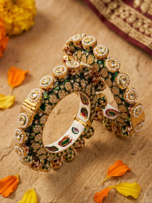 Two ornate Indian Kundan bangles with intricate gold-plated settings, green enamel, and white stones, displayed on a textured beige surface with orange flower petals.