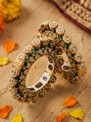 Two ornate Indian Kundan bangles with intricate gold-plated settings, green enamel, and white stones, displayed on a textured beige surface with orange flower petals.