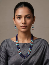A woman wearing a multi-strand beaded necklace and matching drop earrings. The jewelry features dark indigo blue jade beads accented with light blue, pink, and brown beads, styled with a gray saree against a neutral beige background.