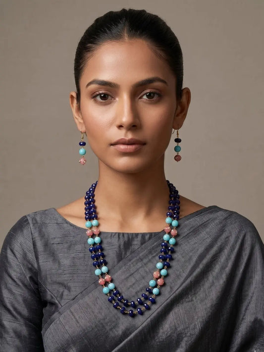 A woman wearing a multi-strand beaded necklace and matching drop earrings. The jewelry features dark indigo blue jade beads accented with light blue, pink, and brown beads, styled with a gray saree against a neutral beige background.