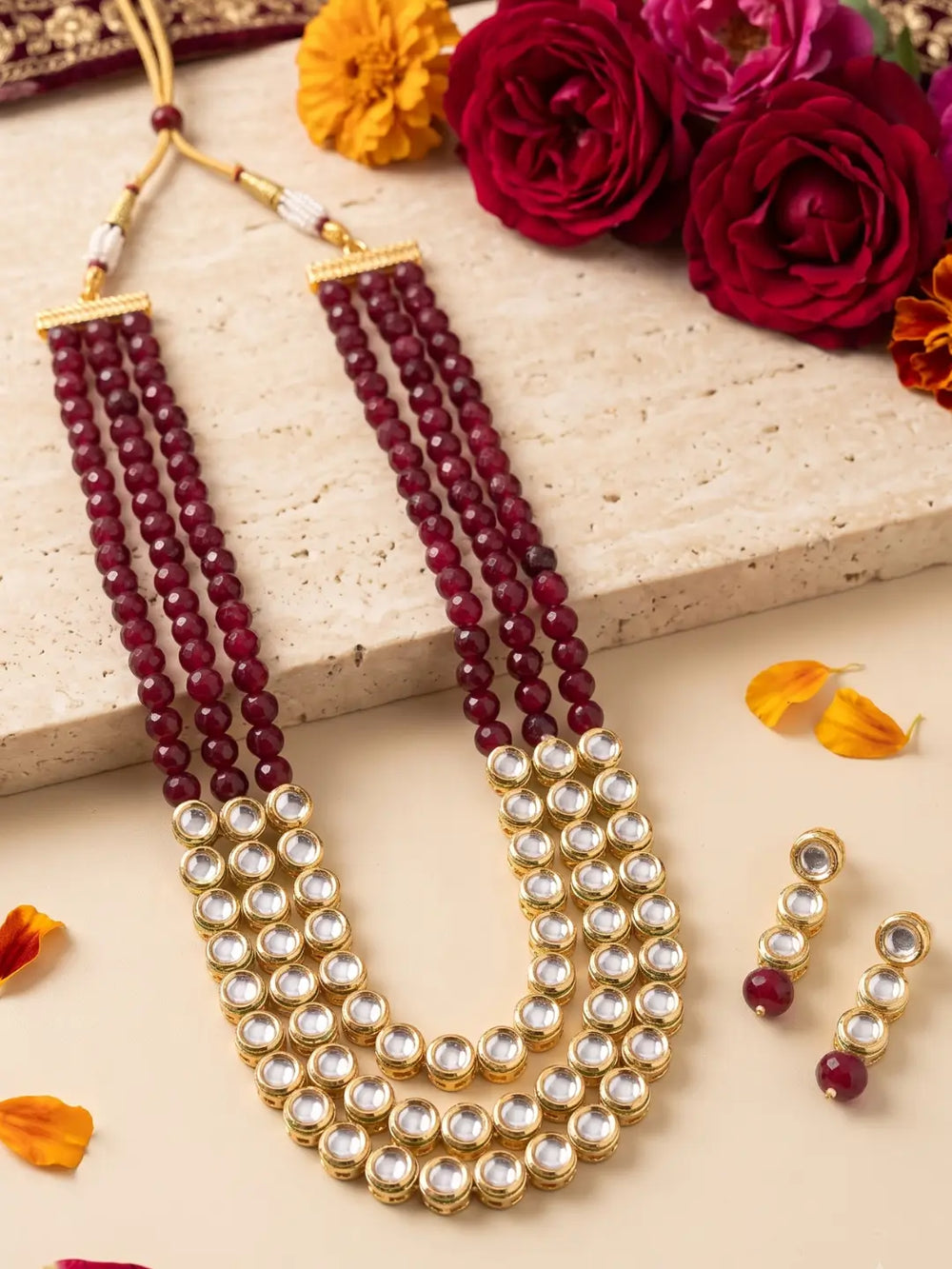 A traditional Indian Kundan jewelry set featuring a multi-strand necklace with deep red beads and gold-plated kundan stones, accompanied by matching drop earrings. The set is styled on a beige stone slab with decorative red roses and orange marigold petals.