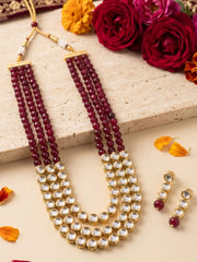 A traditional Indian Kundan jewelry set featuring a multi-strand necklace with deep red beads and gold-plated kundan stones, accompanied by matching drop earrings. The set is styled on a beige stone slab with decorative red roses and orange marigold petals.