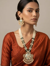A woman wearing an ornate Indian long kundan jewelry set featuring a gold-plated necklace with white beads, green stones, and a large central medallion, paired with matching drop earrings and a rust-colored blouse.