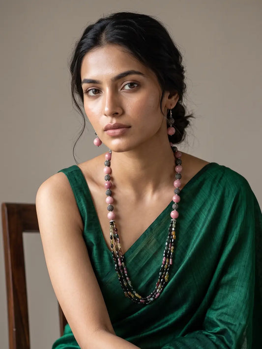 A woman wearing a dark green satin saree and a long multi-strand natural gemstone beaded necklace with pink and dark beads, posed against a neutral beige background.