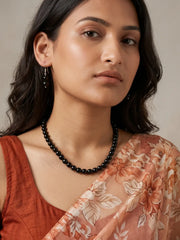 A woman wearing a natural black jade beaded necklace and small black dangling earrings, styled with a rust-colored top and a peach floral dupatta against a neutral beige background.