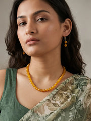 A woman wearing a round yellow beaded necklace and matching drop earrings, paired with a green floral sari against a neutral background.