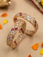 Two ornate gold-plated bangles studded with Polki, CZ and Ruby stones . Displayed on a beige surface with scattered orange and yellow flower petals.