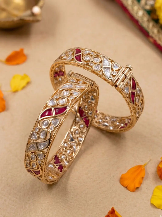 Two ornate gold-plated bangles studded with Polki, CZ and Ruby stones . Displayed on a beige surface with scattered orange and yellow flower petals.