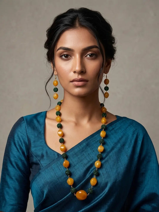 A woman wearing a teal blue sari and matching drop earrings, featuring a yellow and dark green beaded necklace with a large round yellow bead at the center, set against a neutral textured gray background.