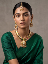 A woman wearing an ornate gold-plated traditional antique kundan necklace with green and red stone accents, matching drop earrings, and a maang tikka, paired with a deep green sari against a neutral gray background.
