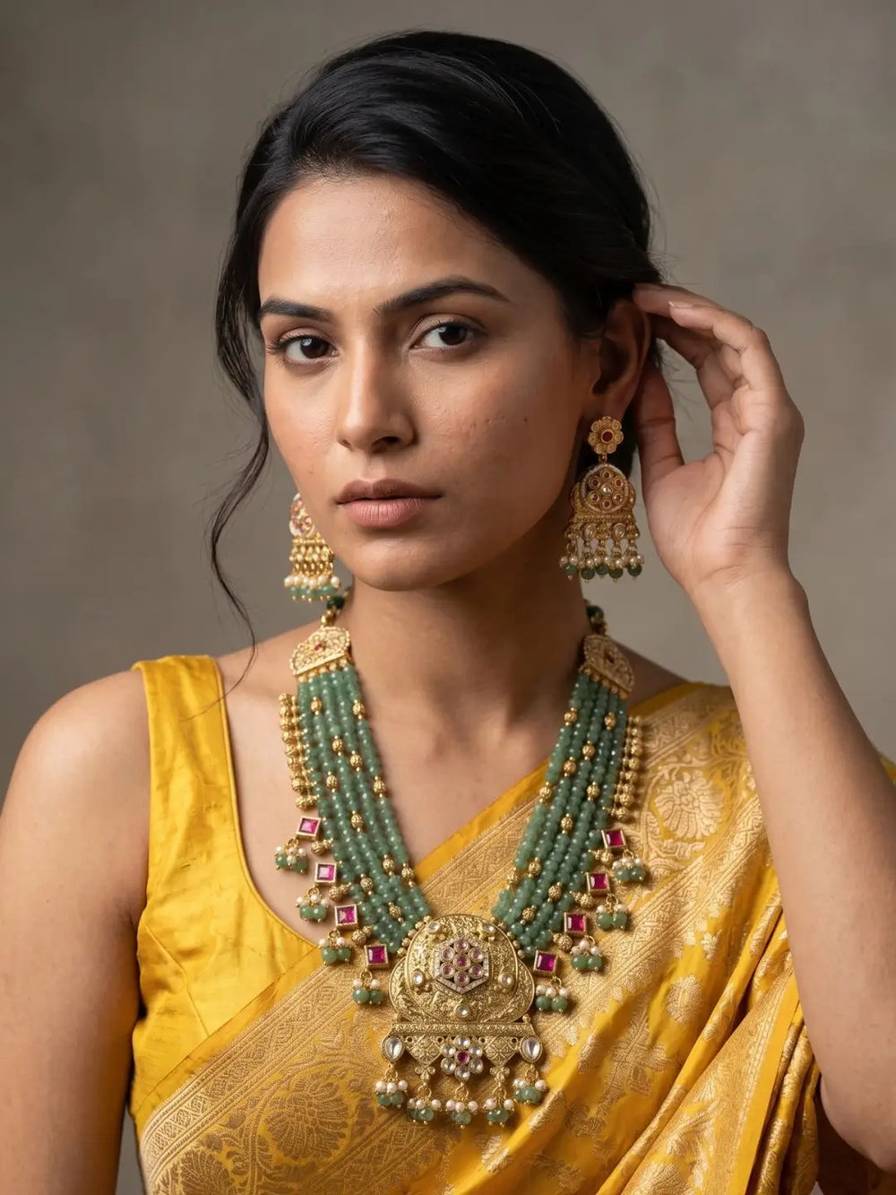 A woman wearing a traditional Indian yellow saree and a large green-and-gold statement necklace with matching earrings, posed against a neutral gray background.