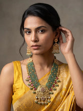 A woman wearing a traditional Indian yellow saree and a large green-and-gold statement necklace with matching earrings, posed against a neutral gray background.