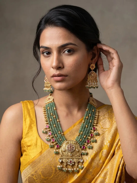 A woman wearing a traditional Indian yellow saree and a large green-and-gold statement necklace with matching earrings, posed against a neutral gray background.