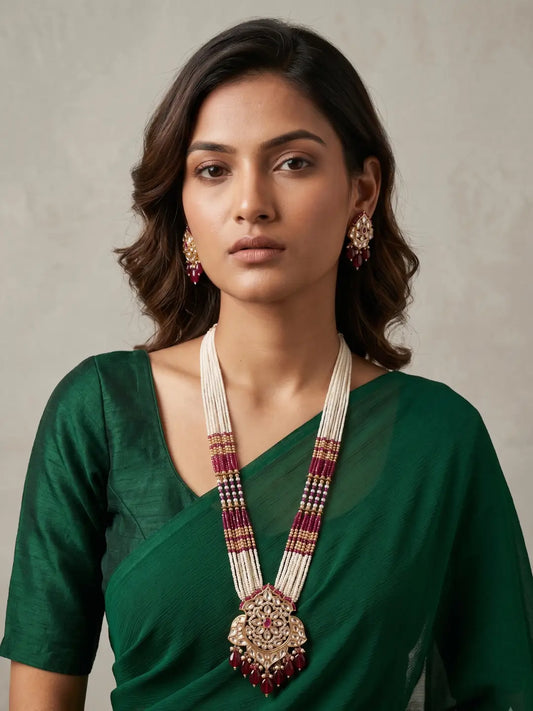 Woman wearing a long red Kundan pearl necklace with multiple pearl-style strands, Red hydro bead sections, an ornate floral pendant and matching traditional earrings.