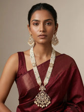 A woman wearing a deep maroon silk saree and a traditional Indian gold-toned long polki necklace with a large ornate pendant, paired with matching earrings, set against a plain beige background.