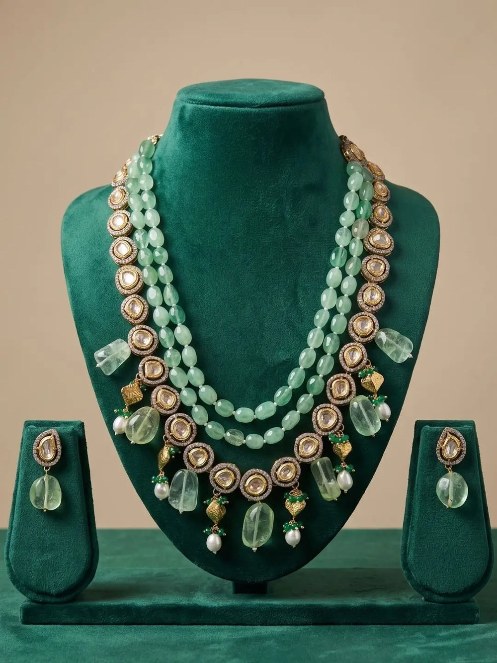 A three-strand statement necklace set displayed on a dark green velvet jewelry bust, accompanied by matching drop earrings on display stands. The necklace features light green beads, gold-toned circular medallions, clear faceted beads, and small white pearl accents, set against a solid beige background.