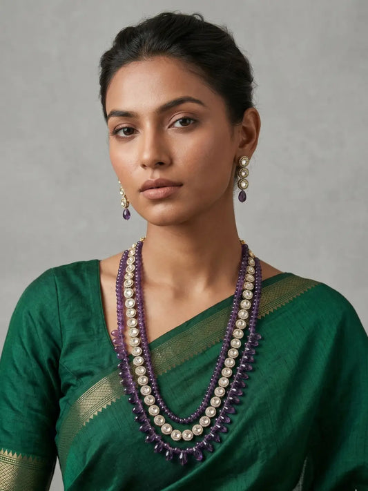 A woman wearing a dark green saree with a gold border and a multi-strand statement necklace featuring purple gemstone amethyst beads and kundan accents, set against a neutral gray background.
