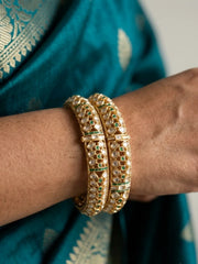 A close-up of a person's wrist wearing two stacked gold bangles, polki kundan bangles adorned with white stones and small green accents, against a teal sari background.