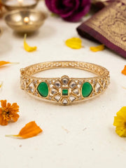 Gold-plated bangle bracelet with green stone inlays and clear mirror-like stones, displayed on a white surface with decorative flowers and traditional props.
