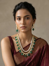 A woman wearing a traditional Indian outfit with a deep maroon saree and a multi-strand Kundan, pearl, and green stone necklace, along with matching drop earrings, set against a neutral beige background.