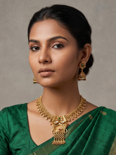 A woman wearing an ornate gold-toned traditional necklace with a large pendant and matching dangling earrings, paired with a green sari against a neutral gray studio background.