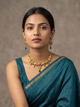A woman wearing a teal blue saree with a gold border, styled with a gold plate antique golden necklace featuring pink stones and green bead drops, and matching gold earrings, set against a neutral textured brown background.