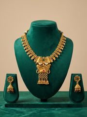 Gold-toned traditional Indian jewelry set displayed on a dark green velvet stand, featuring a heavy necklace with a central peacock pendant and matching drop earrings.