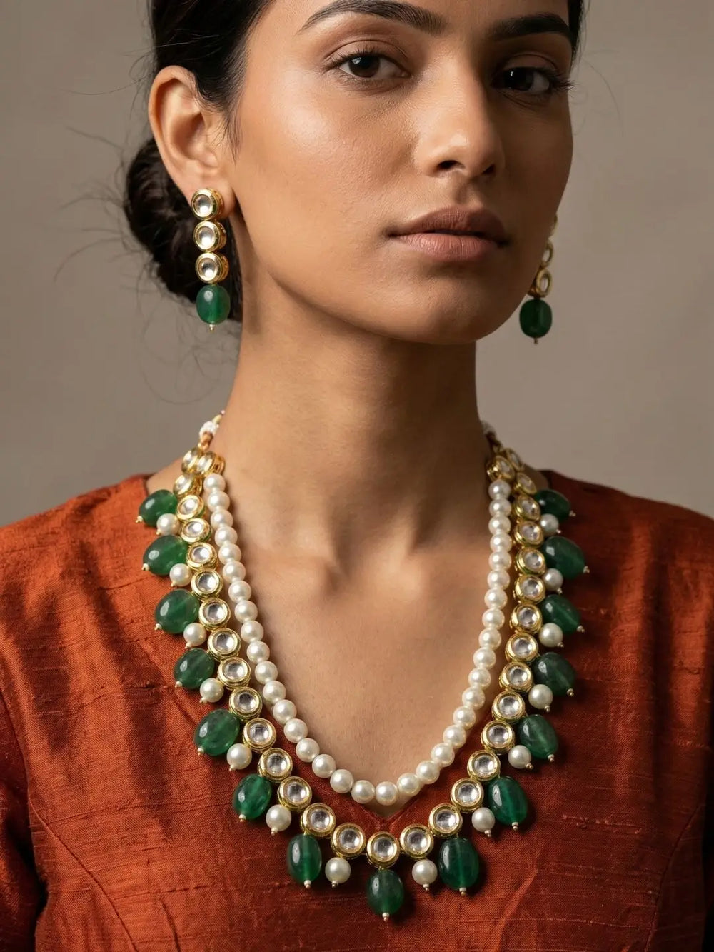 A close-up of a woman wearing a traditional Indian kundan jewelry set featuring a double-strand pearl necklace with green and gold plated kundan string and matching drop earrings, and a rust-orange textured top.