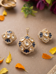 Gold-tone Indian Kundan jewelry set featuring a matching pendant and two matching earrings, decorated with clear stones, white crystals, and navy blue enamel details, displayed on a textured beige surface with scattered orange and yellow flower petals.