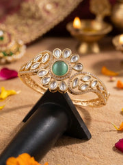 Gold-plated kundan bracelet with green gemstone on a black stand, with Diwali decorations in the background.