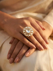 Close-up of a hand wearing an ornate gold-plated kundan ring with a large central gemstone, set against a soft-focus background.