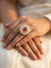 A close-up of a person's hands resting on cream-colored fabric, wearing a gold-plated red kundan ring with a central round red gemstone surrounded by small clear polki stones.