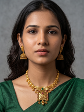 A woman wearing a gold-toned traditional antique gold necklace with a central green and red stone pendant, matching dangling earrings, and a green blouse, photographed against a neutral gray background.