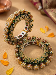 Two gold-plated Indian Kundan bangles displayed on a textured beige surface with scattered yellow flower petals. The bangles feature intricate green enamel work with white pearls and kundan stones