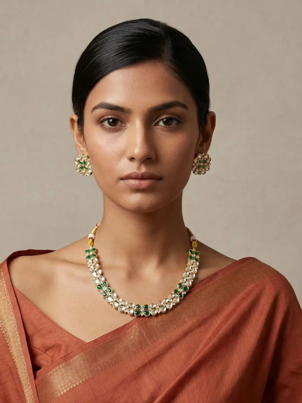 A woman wearing a gold-plated kundan necklace and matching earrings with green and white stones, paired with a rust-colored sari, against a neutral beige background.
