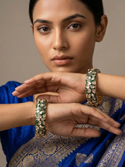 A close-up of a woman wearing ornate gold bangles with green and white floral meenakari paired with a royal blue sari featuring intricate gold zari work.