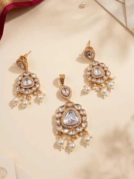 Gold-plated traditional Indian kundan pendant earrings and matching pendant with white faux pearls and clear stones, arranged on a beige satin fabric background.