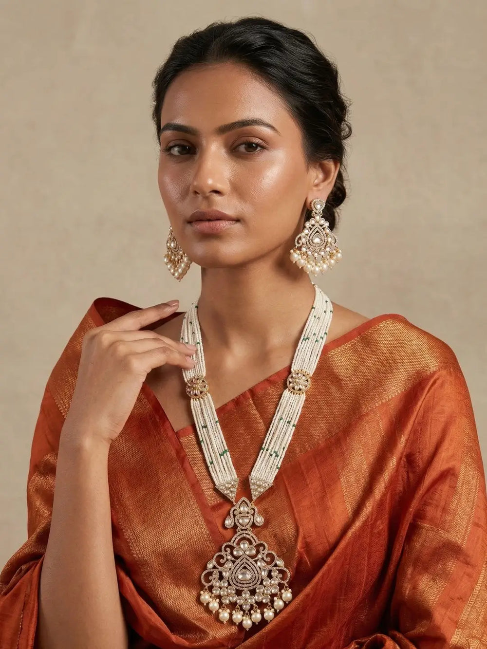 A woman wearing a traditional Indian outfit in rust-orange silk, with a large white beaded long polki necklace and matching earrings with gold-toned details, posed against a neutral beige background.