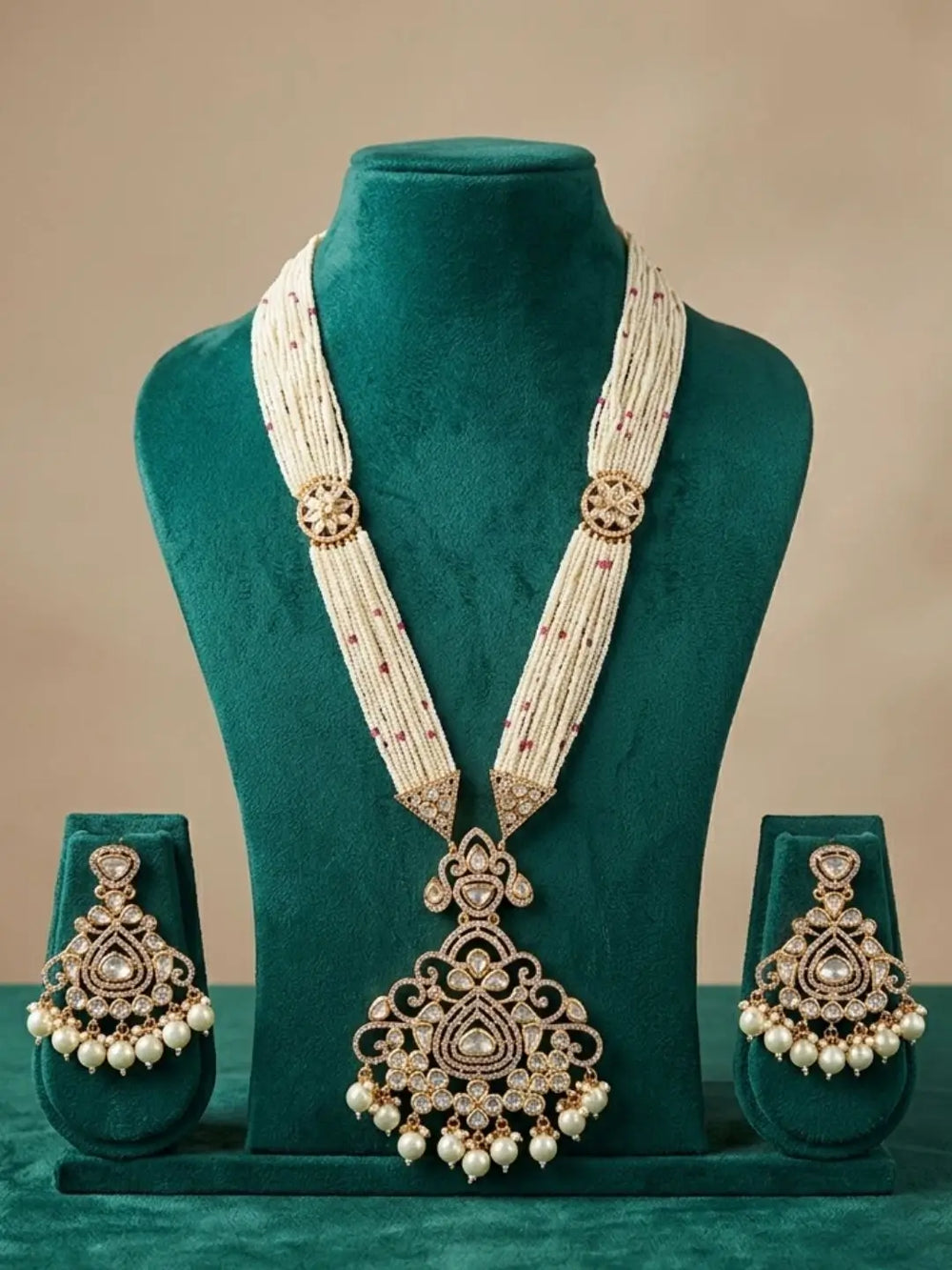 A traditional Indian jewelry set displayed on a dark green velvet mannequin bust. The set includes a long necklace with a large ornate pendant, matching drop earrings, and a pair of matching chain necklaces, all featuring gold-toned metalwork, white pearls, and small red stone accents.