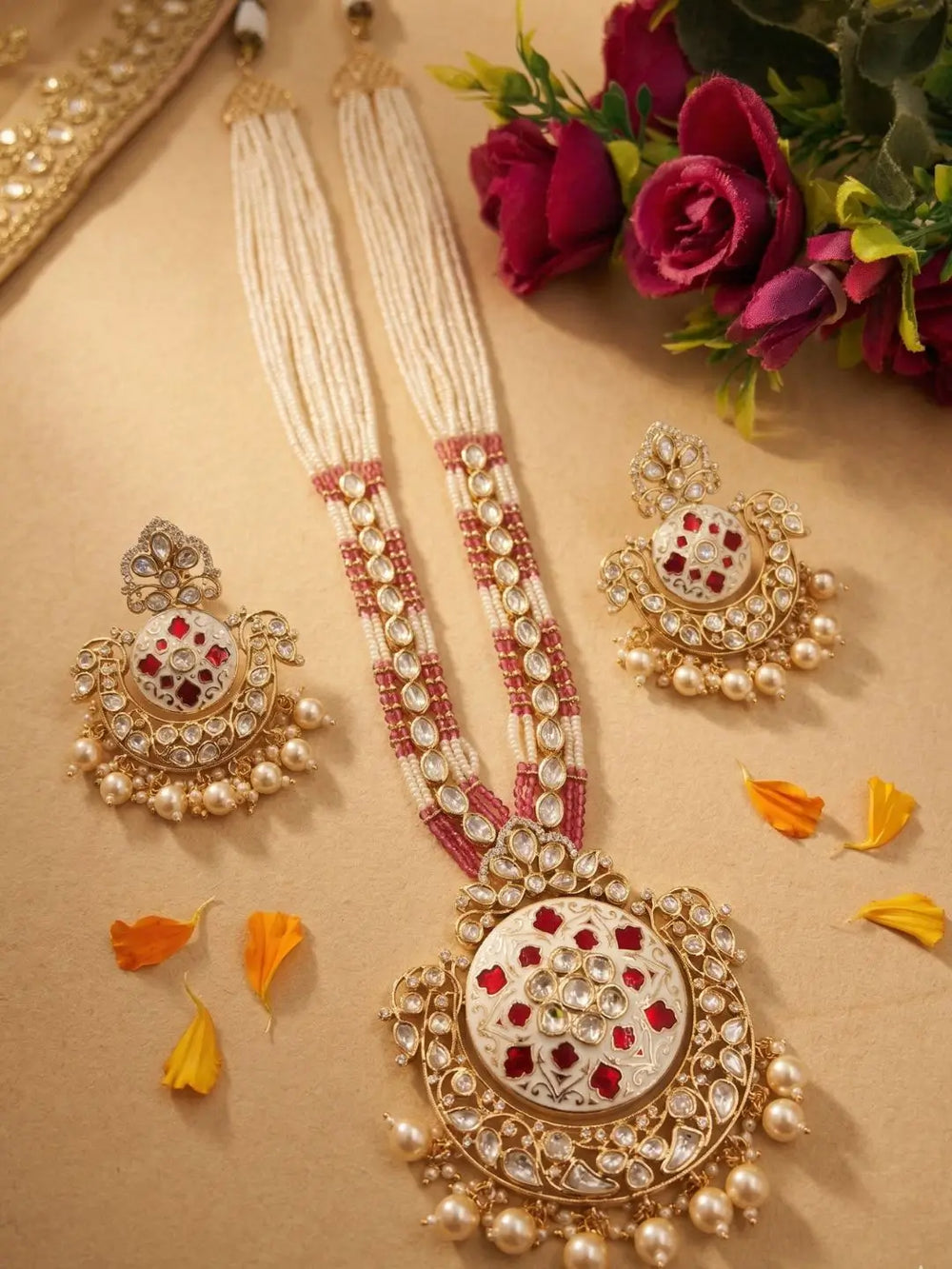An ornate Indian long bridal jewelry set displayed on a beige surface, featuring a long necklace with a large circular pendant, matching earrings, and pearl strands, decorated with gold, white stones, and red accents, styled with flowers and orange petals.