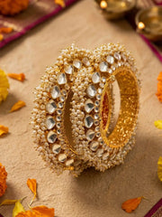 A stack of ornate gold-plated paachi kundan jadau kada bangles decorated with white pearls, arranged on a textured beige surface with scattered orange flower petals.